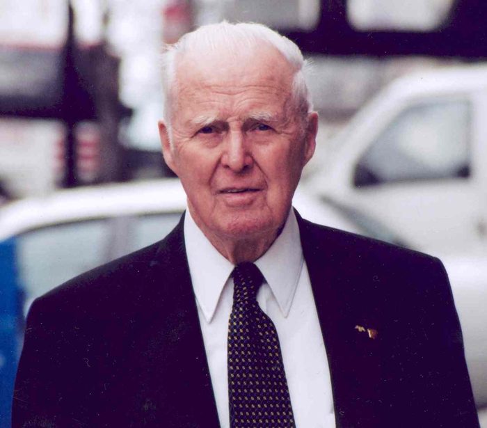 Norman Borlaug at 90, white-haired, in a dark suit and dotted tie on a city street