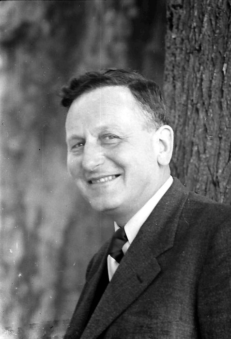Photograph of Paul Müller smiling, in a dark suit and tie, in front of a tree trunk