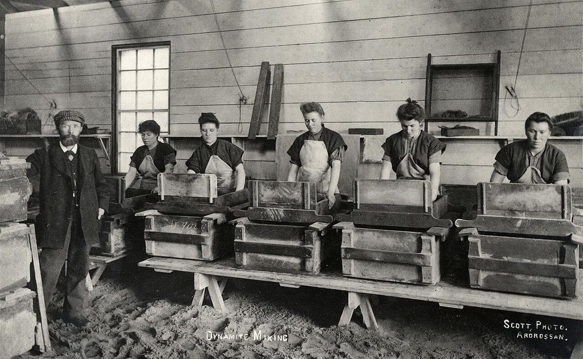 Women in aprons mixing dynamite in wooden boxes along a bench, with a man standing at the left, inside a timber factory shed