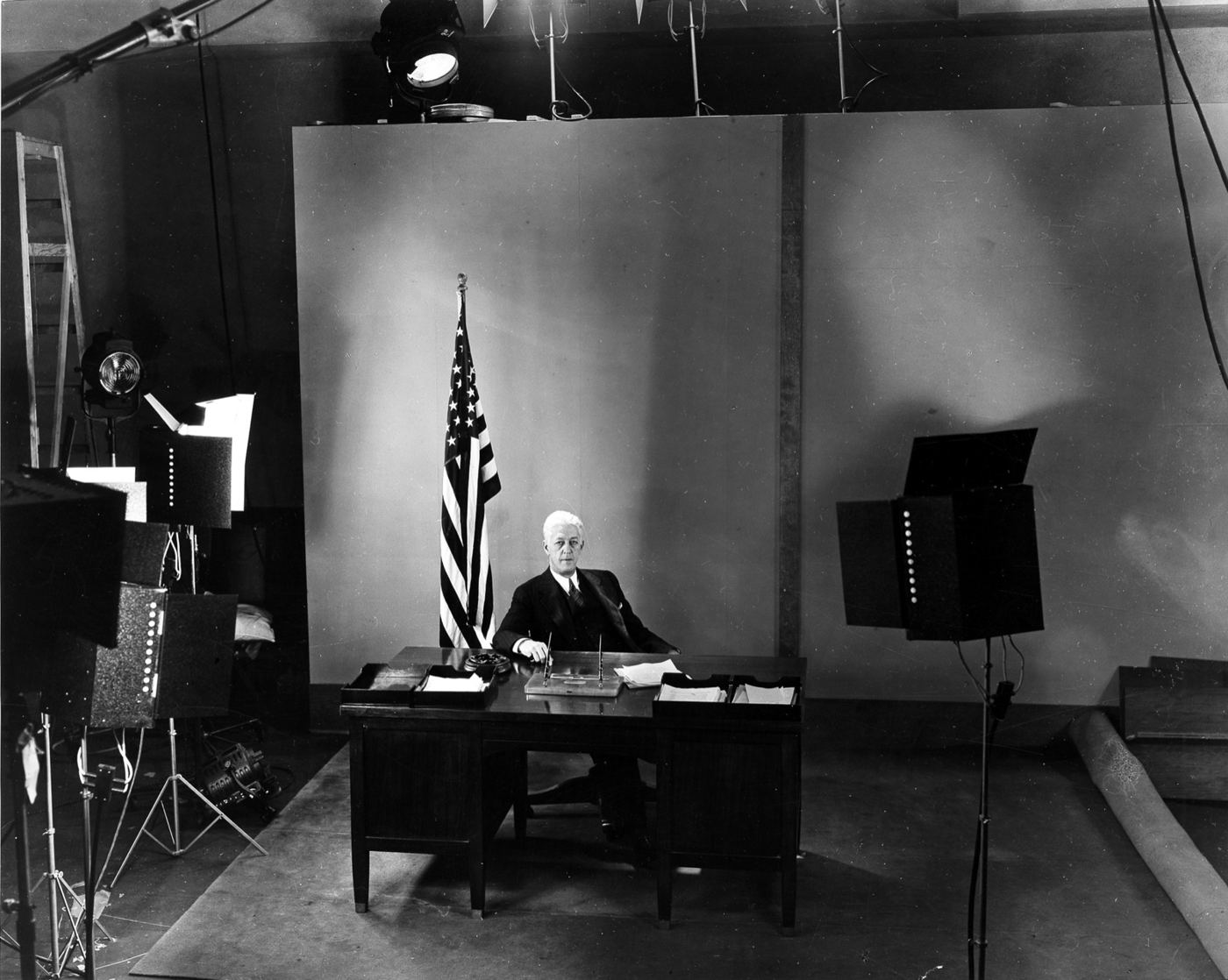 Ferdinand Silcox, white-haired, in a dark suit, seated at a desk beside a US flag in a studio surrounded by lights